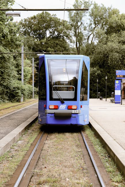 The image depicts a modern yellow and grey tram approaching a stop on a railway track, set amidst a green outdoor environment. The tram is sleek, with large windows and a streamlined design, contrasti