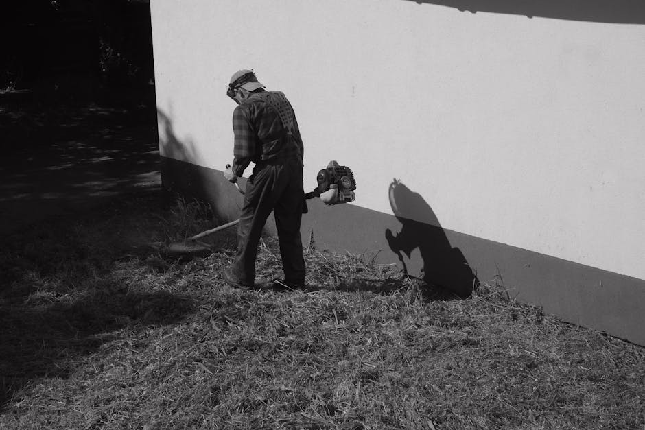 A man engaged in garden maintenance outdoors, wearing a plaid shirt, trousers, and a hat, operating a handheld blower or leaf vacuum while working near a garden bed with overgrown grass and soil. The 